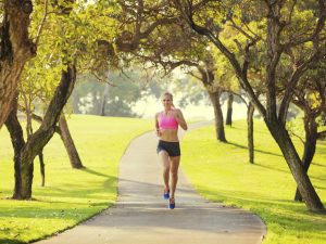 Woman in a pink sports top and black shorts running on a paved park path under leafy trees, sunlight filtering through branches.