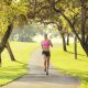 Woman in a pink sports top and black shorts running on a paved park path under leafy trees sunlight filtering through branches