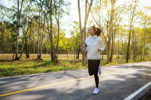 Woman jogging on a tree lined park road in a white jacket and black leggings listening to earbuds on a sunny day